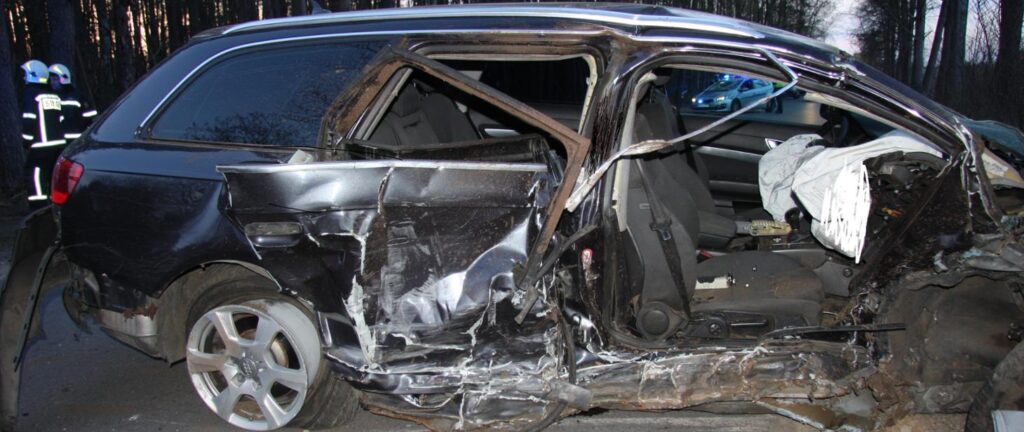 Rozwieziony samochód osobowy po wypadku drogowym, w tle strażacy w mundurach oraz policyjny radiowóz.