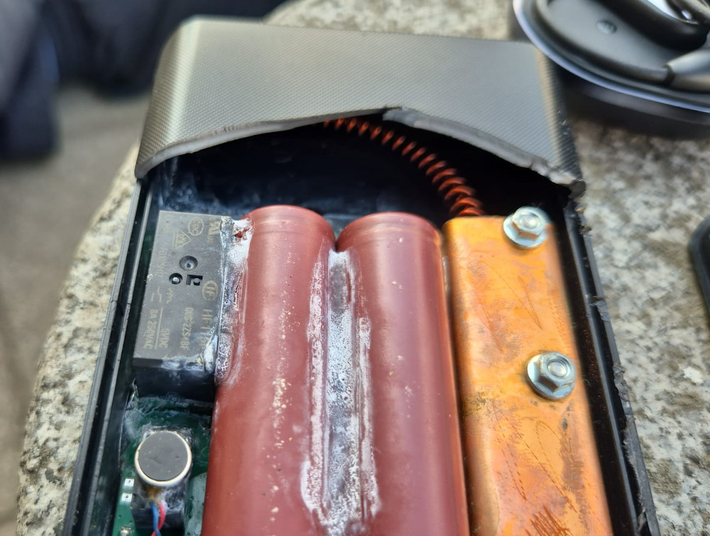 Uszkodzony akumulator z widocznymi elementami elektronicznymi, w tym czerwonymi ogniwami i metalowymi złączami.