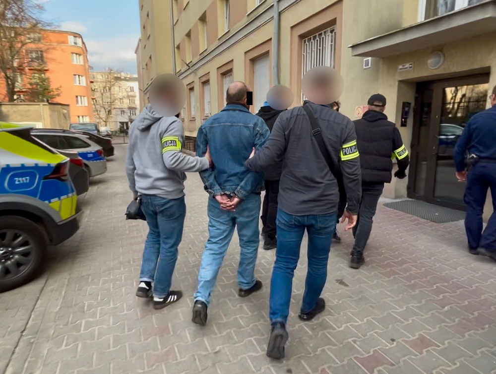 Mężczyzna w dżinsowej kurtce prowadzony przez policjantów na chodniku obok radiowozu.