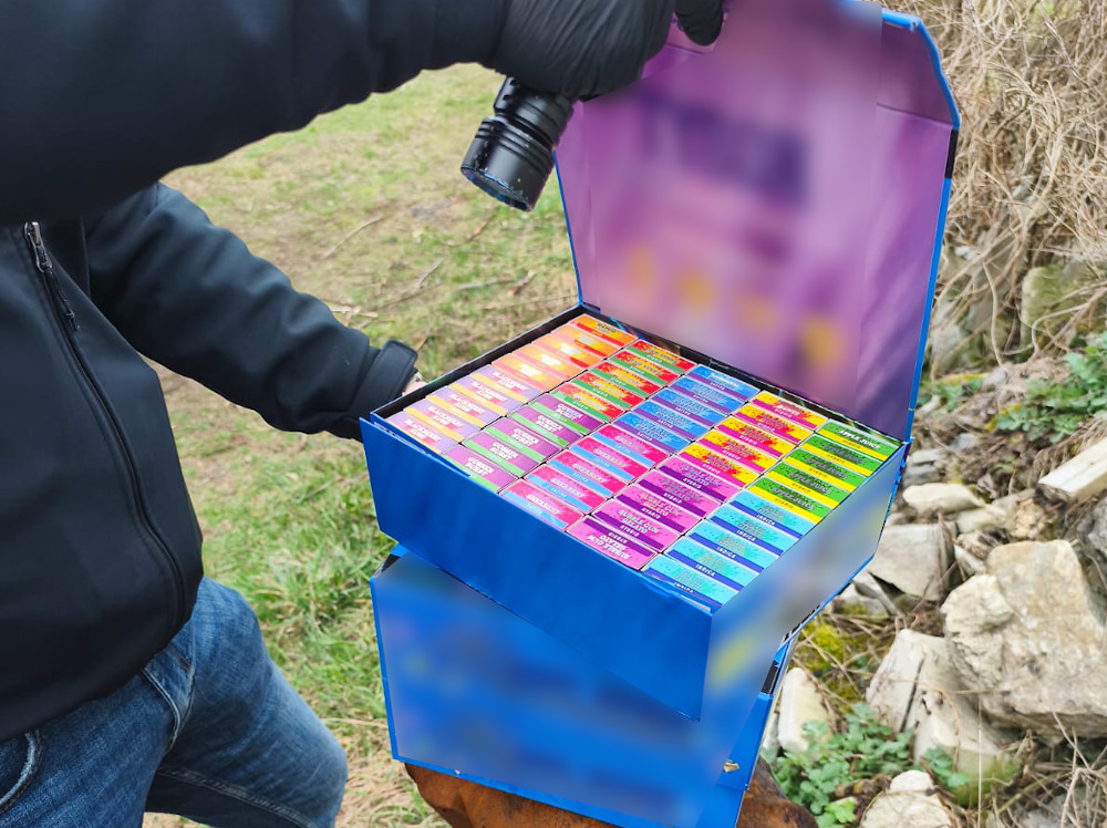 Mężczyzna w czarnej kurtce trzyma otwartą niebieską skrzynkę z kolorowymi opakowaniami.