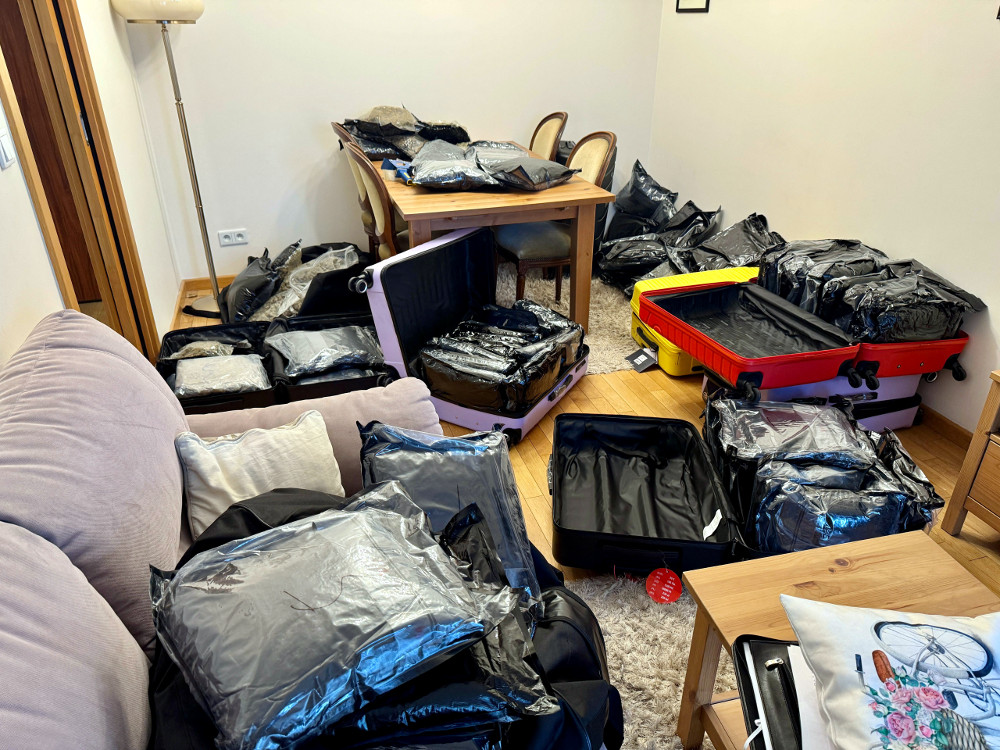 W pomieszczeniu mieszkalnym rozłożone są torby i walizki w czarnych workach, zajmujące całą przestrzeń.