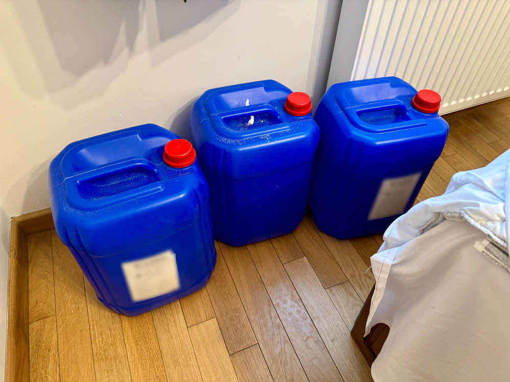Trzy niebieskie pojemniki z czerwoną zakrętką stoją obok łóżka na drewnianej podłodze.