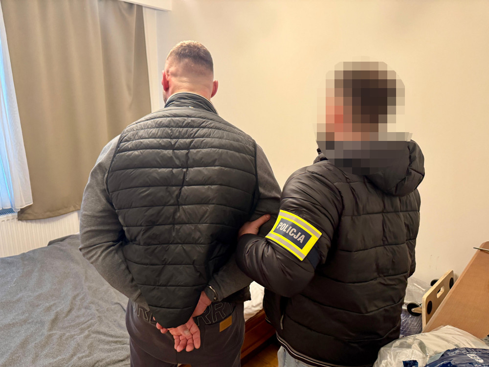 Mężczyzna w szarej kurtce jest zatrzymywany przez policjanta w czarnej kurtce w pokoju z łóżkiem.