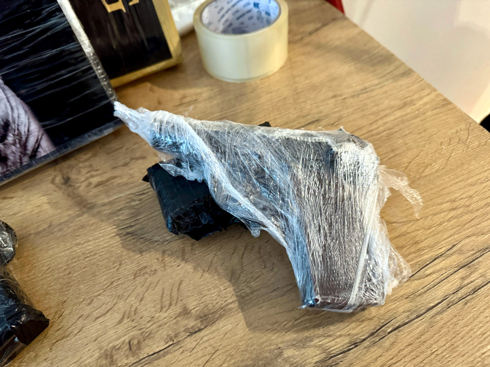 Pistolet owinięty folią stretch leży na drewnianym stole obok czarnych pakunków i taśmy klejącej.