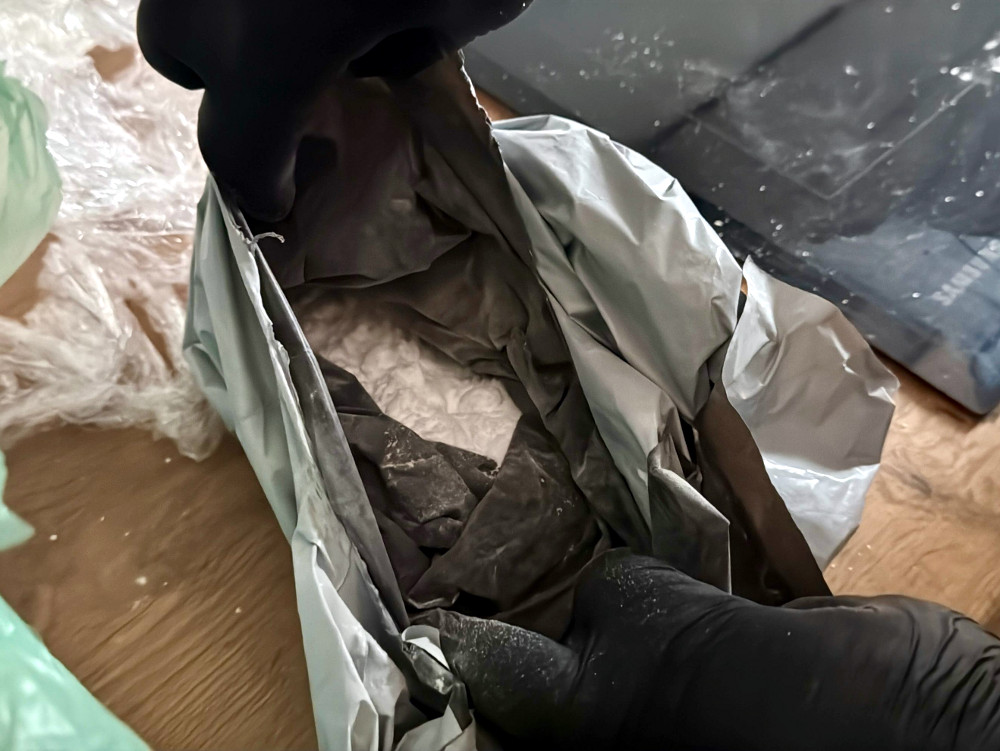 Osoba w czarnych rękawiczkach otwiera szczelnie zamknięte opakowanie, w którym znajduje się biały proszek.