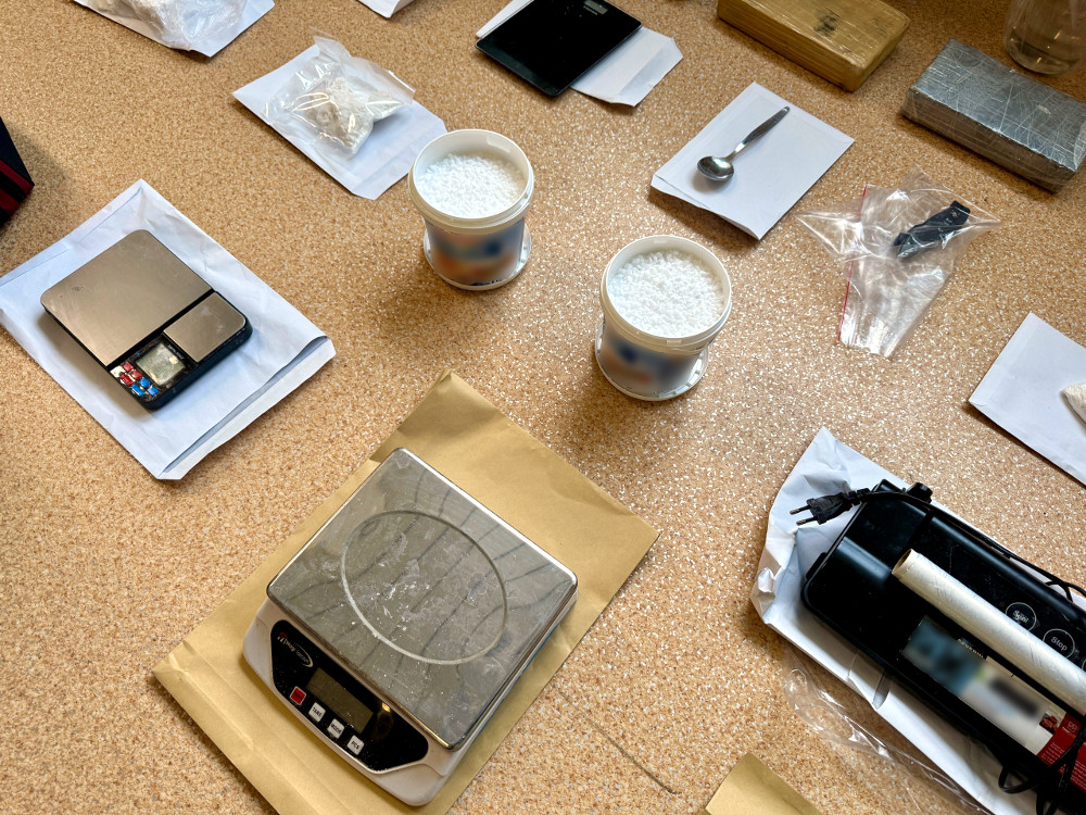 Na blacie widoczne są wagi elektroniczne, pojemniki z białym proszkiem oraz inne akcesoria, w tym łyżka i telefon.