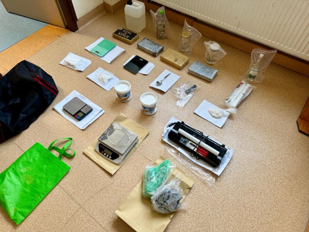 Na podłodze leżą różne przedmioty, w tym wagi, opakowania z białym proszkiem, telefon i torba.