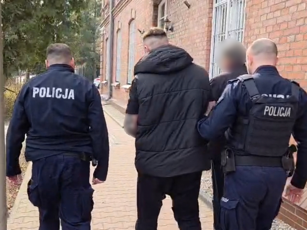 Trzech policjantów eskortuje zatrzymanego mężczyznę w czarnej kurtce wzdłuż chodnika obok ceglanego budynku.