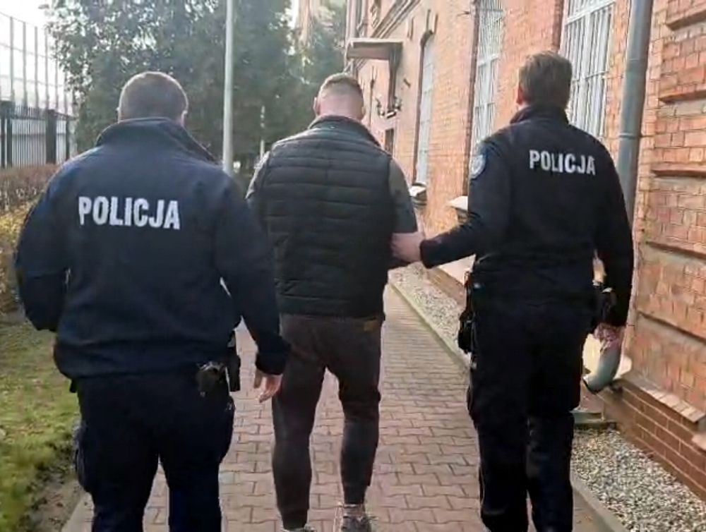 Mężczyzna prowadzony przez dwóch policjantów w mundurach, idący chodnikiem wzdłuż budynku.