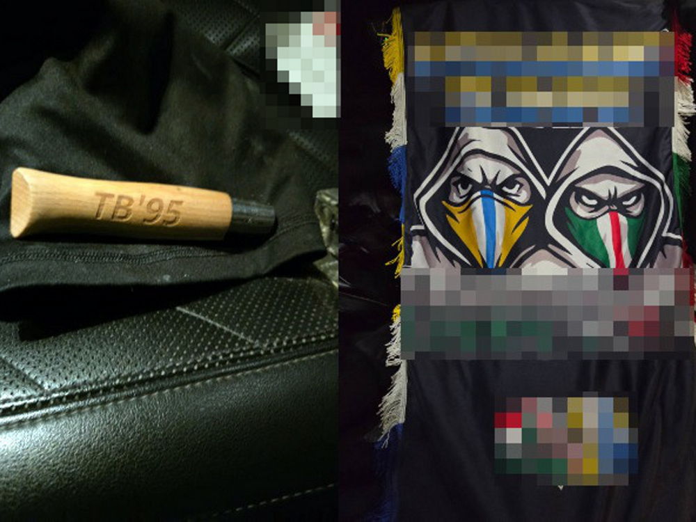 Drewniana pałka z wygrawerowanym napisem "TB'95" leży na czarnym tle, obok kolorowy szalik z grafiką dwóch postaci w kapturach.