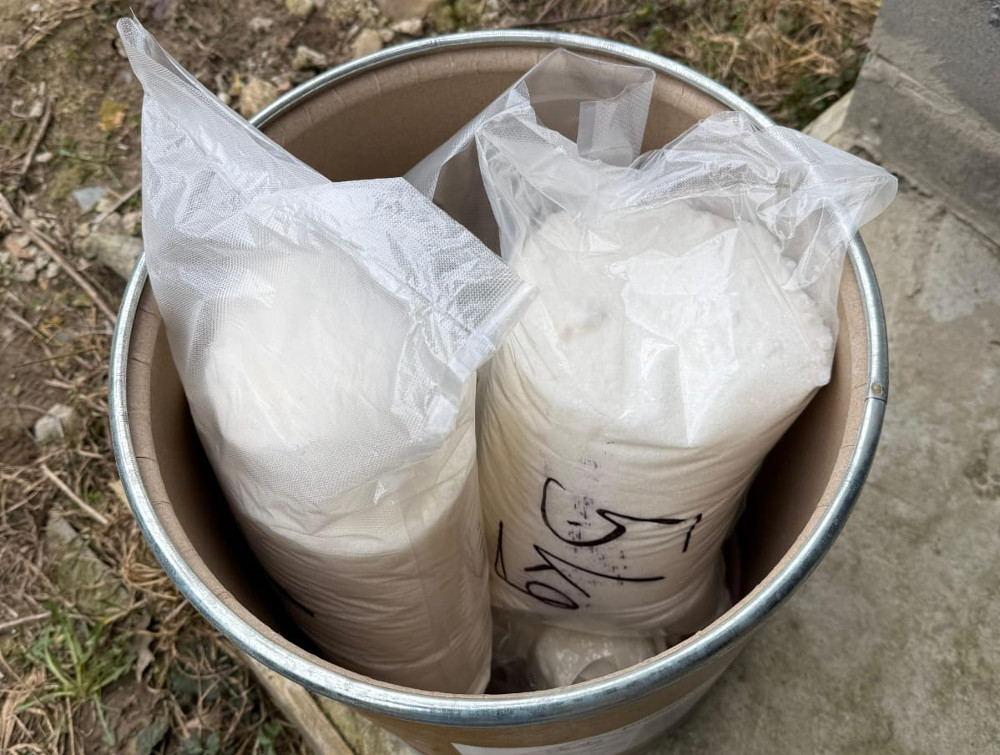 Worki z białym proszkiem umieszczone w metalowej beczce, otoczone kamieniami i trawą.