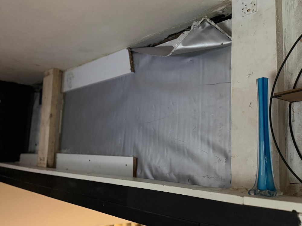 Uszkodzona ściana z odsłoniętym fragmentem, obok szklana waza na półce.