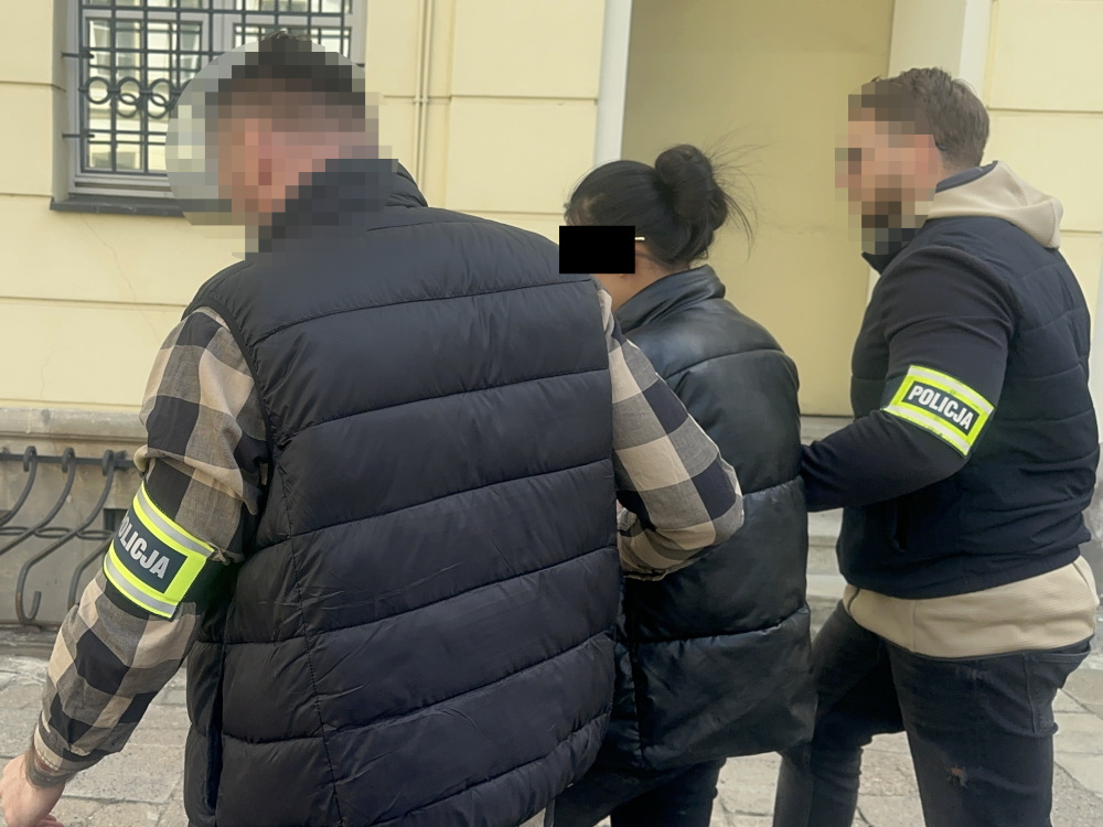 Grupa policjantów w odzieży roboczej prowadzi osobę w czarnej kurtce na zewnątrz budynku.