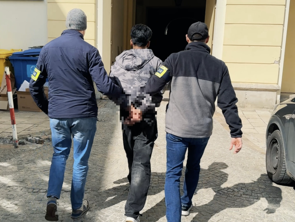 Dwaj mężczyźni w mundurach prowadzą zatrzymanego mężczyznę w szarym dresie na ulicy obok budynku.