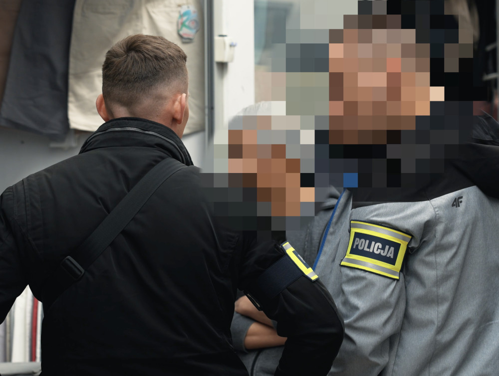Mężczyzna w czarnej kurtce rozmawia z funkcjonariuszami policji w miejskim otoczeniu.