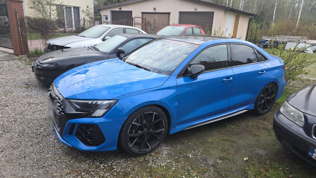 Niebieski samochód sportowy zaparkowany na podjeździe wśród innych pojazdów. W tle widać budynek mieszkalny.