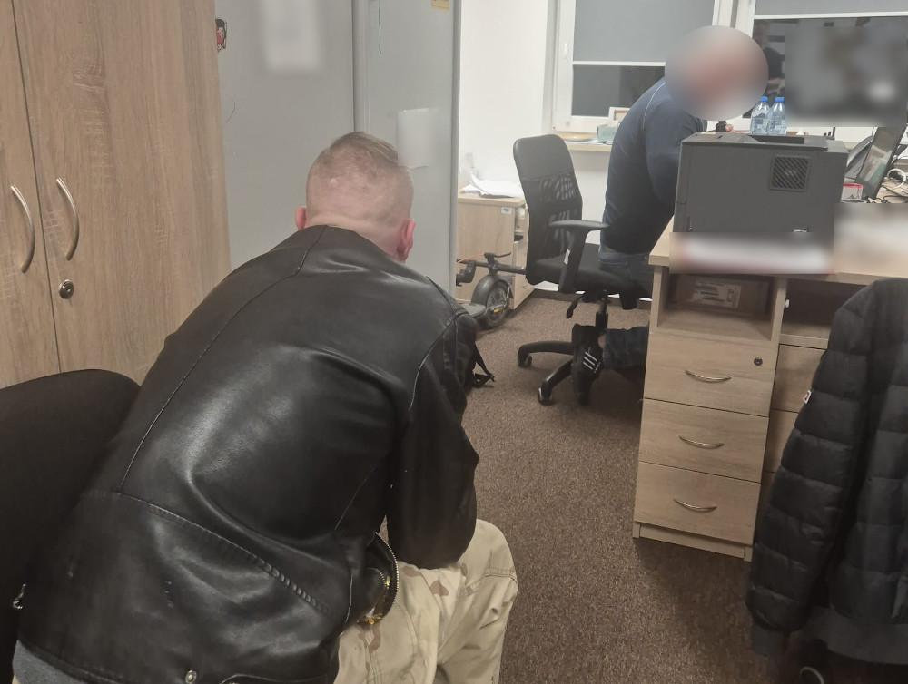 Mężczyzna w skórzanej kurtce siedzący na krześle, obserwujący drugiego mężczyznę przy biurku w biurze.