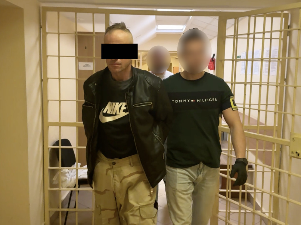 Mężczyzna w skórzanej kurtce prowadzony przez policjanta w mundurze w korytarzu budynku.