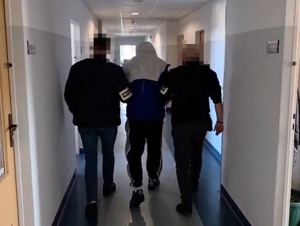 Mężczyzna prowadzony przez dwóch funkcjonariuszy w korytarzu budynku.