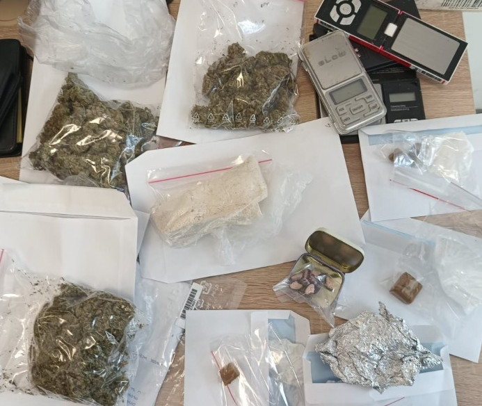 Na stole rozłożone są różne opakowania z substancjami, w tym marihuaną, białym proszkiem i tabletkami. Obok znajduje się waga elektroniczna.
