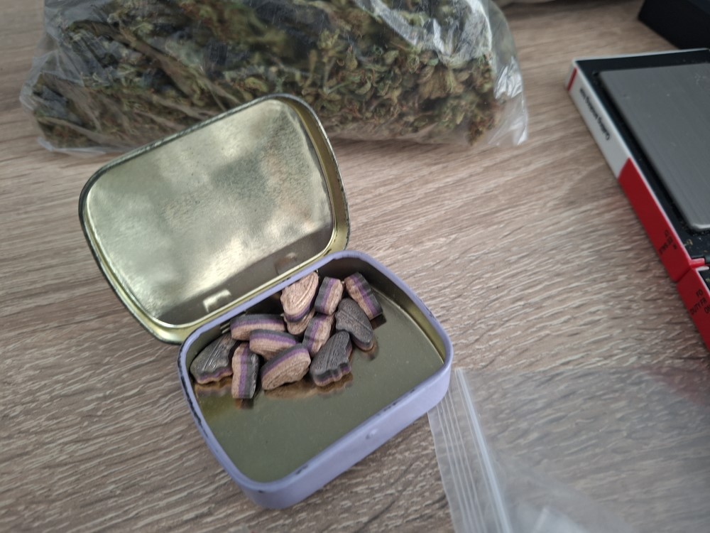 Otwarta puszka z kolorowymi tabletkami leży na stole obok torebki z suszem roślinnym.