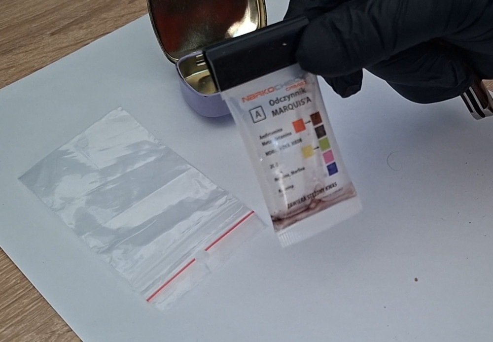 Ręka w czarnej rękawiczce trzyma odczynnik narkotykowy obok pustej przezroczystej torebki na tle białego papieru.