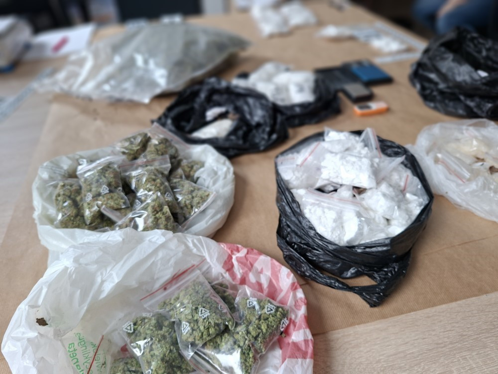 Na stole rozłożone są pakunki z substancjami, w tym torebki z marihuaną i białym proszkiem, w różnych torbach.