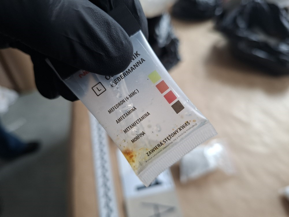 Osoba w czarnych rękawiczkach trzyma opakowanie z testem narkotykowym, na którym widnieją nazwy substancji, takich jak mefedron i amfetamina.