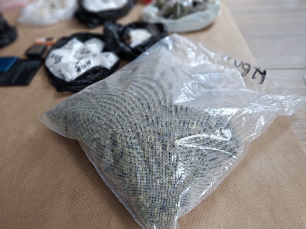 Zamknięta w przezroczystej torbie foliowej roślinna substancja leży na brązowym podłożu obok innych pakunków.