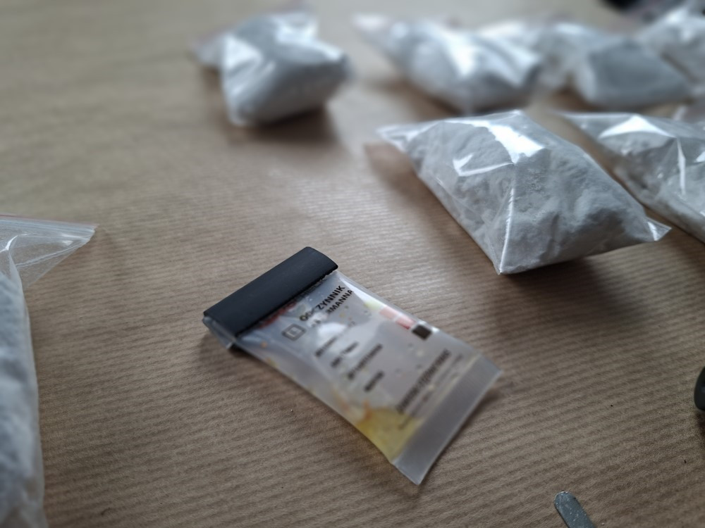Sześć torebek z białym proszkiem leży na brązowym stole obok przezroczystej saszetki z etykietą.