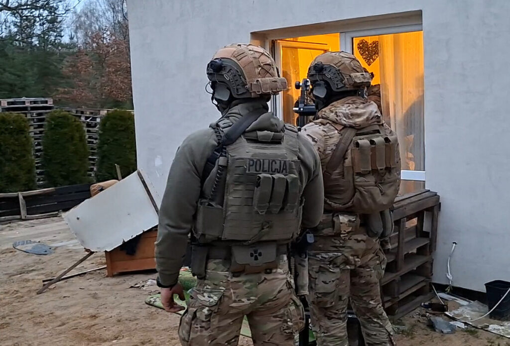 Dwaj policjanci w maseczkach i kamizelkach taktycznych stoją przed domem, obserwując wnętrze przez okno.