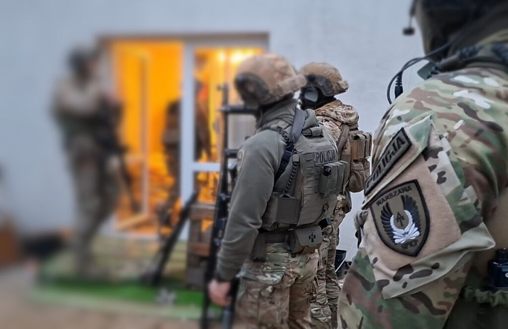 Grupa uzbrojonych funkcjonariuszy policji przygotowuje się do akcji w pobliżu otwartych drzwi budynku.