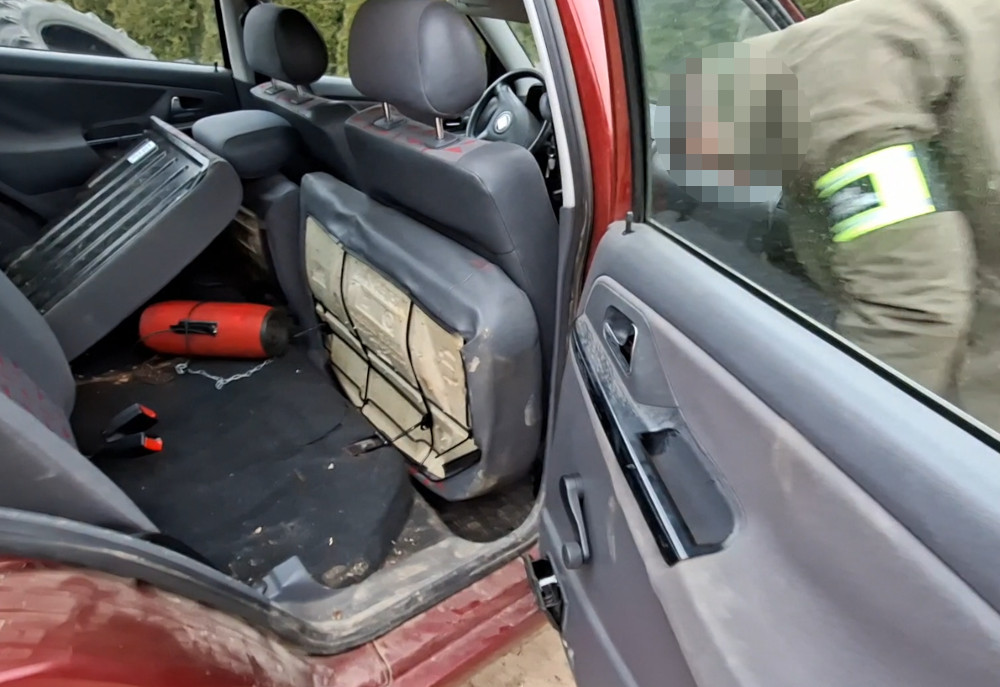 Wnętrze czerwonego samochodu z przewróconym fotelem, na podłodze leży czerwona butla. Obok mężczyzna w mundurze przeszukuje auto.