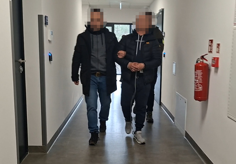 Mężczyzna w dresie prowadzony przez policjanta korytarzem w budynku administracyjnym. W tle widoczne drzwi i gaśnica.