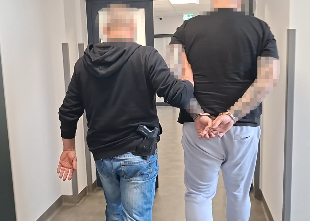 Funkcjonariusz w czarnej bluzie prowadzi aresztowanego mężczyznę z rękami związanymi za plecami korytarzem w budynku.