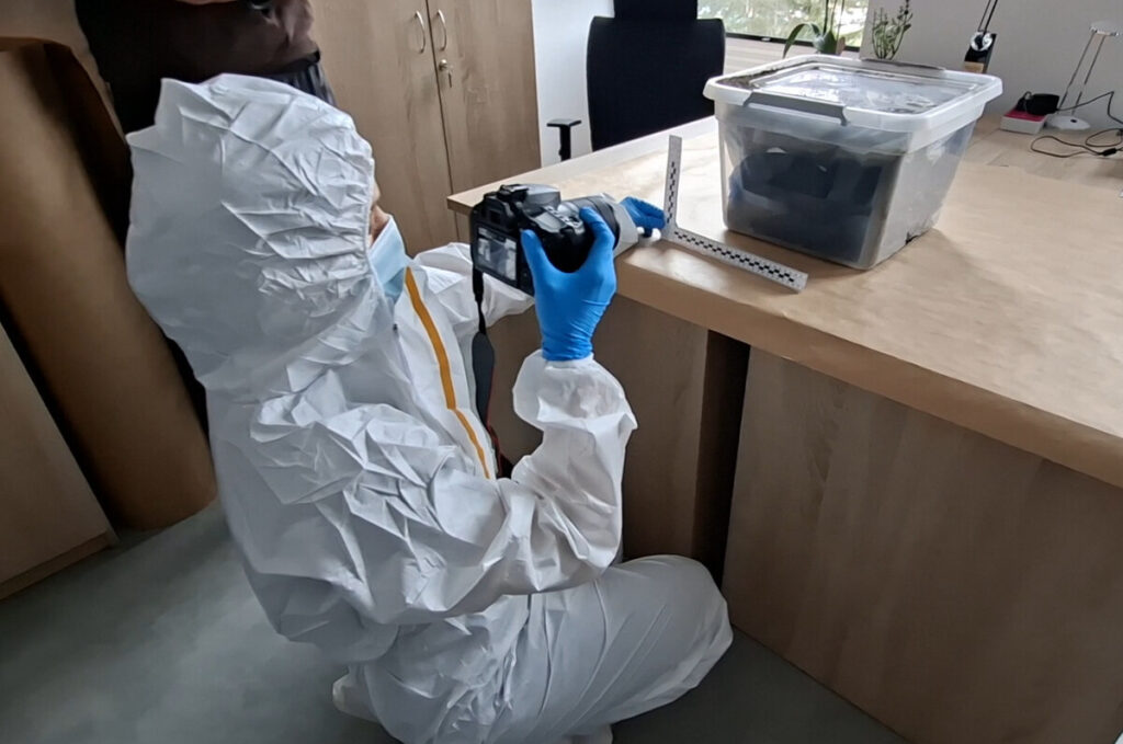 Osoba w białym kombinezonie ochronnym robi zdjęcie aparatem fotograficznym przy biurku z przezroczystym pojemnikiem.