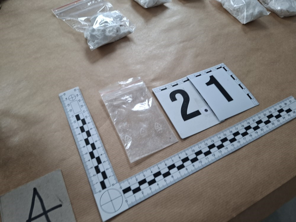 Pakiety z białym proszkiem na brązowym papierze, obok znaki identyfikacyjne i linijka.