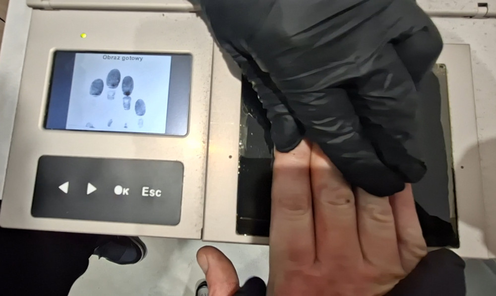 Osoba w czarnych rękawiczkach skanuje odcisk palca na urządzeniu z wyświetlaczem, na którym widoczne są gotowe odciski.