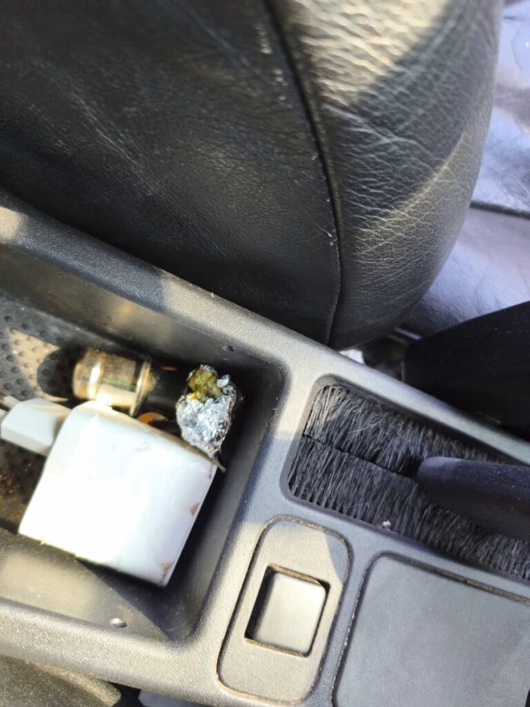 Czarna tapicerka fotela samochodowego z widocznymi przedmiotami w schowku, w tym folią aluminiową i ładowarką.