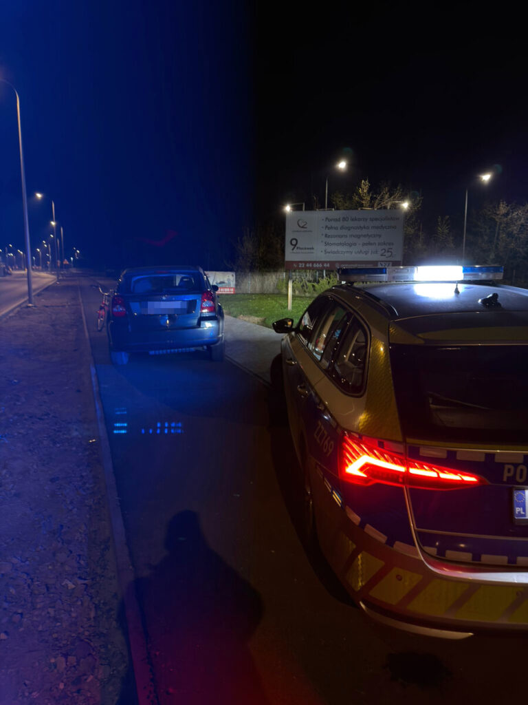 Policyjny radiowóz z włączonymi światłami sygnalizacyjnymi zatrzymany obok osobowego samochodu na nocnej ulicy.
