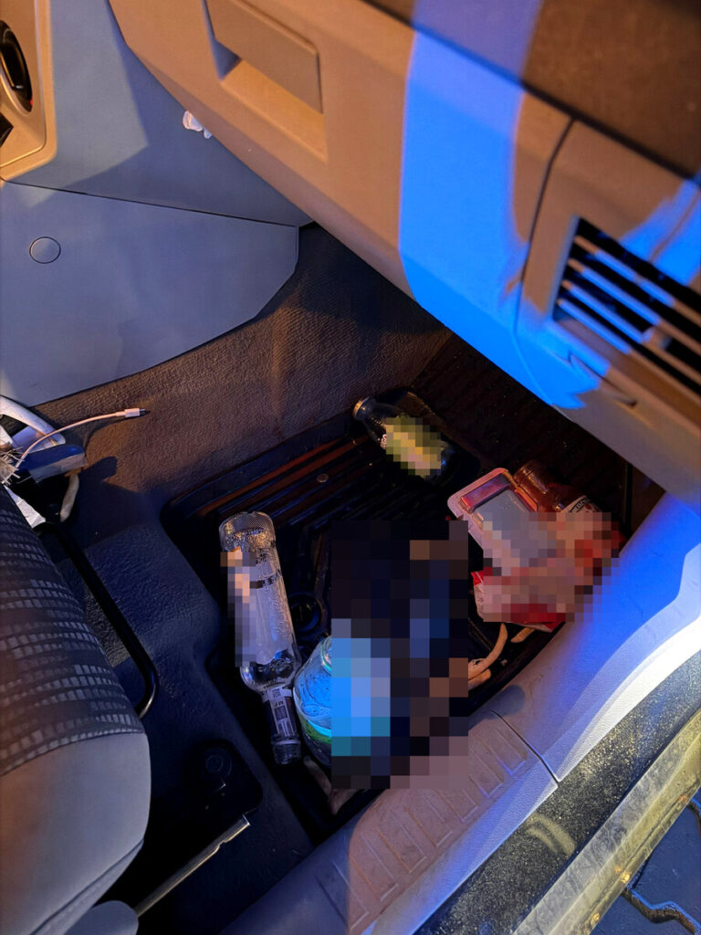 Na podłodze samochodu widoczne są różne butelki, w tym przezroczysta butelka po alkoholu oraz napoju, obok leży telefon.
