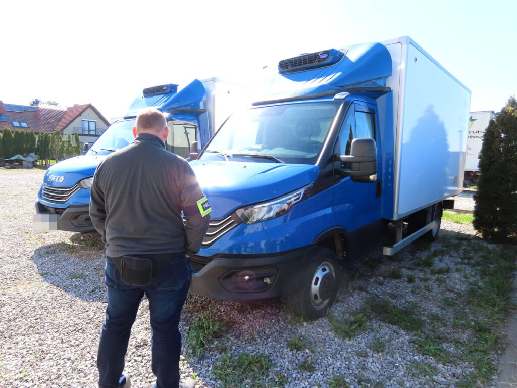Mężczyzna w mundurze policyjnym przegląda niebieskie samochody dostawcze na terenie parkingu.