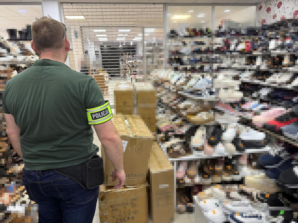 Mężczyzna w mundurze z opaską "POLICJA" obserwujący zmagazynowane paczki w sklepie obuwniczym.