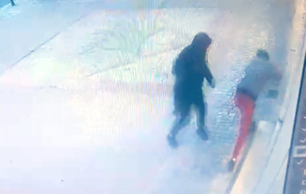 Dwie osoby na ulicy, jedna w czarnym płaszczu, druga w szarej bluzie i czerwonych spodniach; jedna z nich nachyla się w stronę ściany.