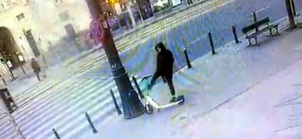 Mężczyzna w czarnej kurtce kopie elektryczną hulajnogę na pustej ulicy w miejskim centrum.