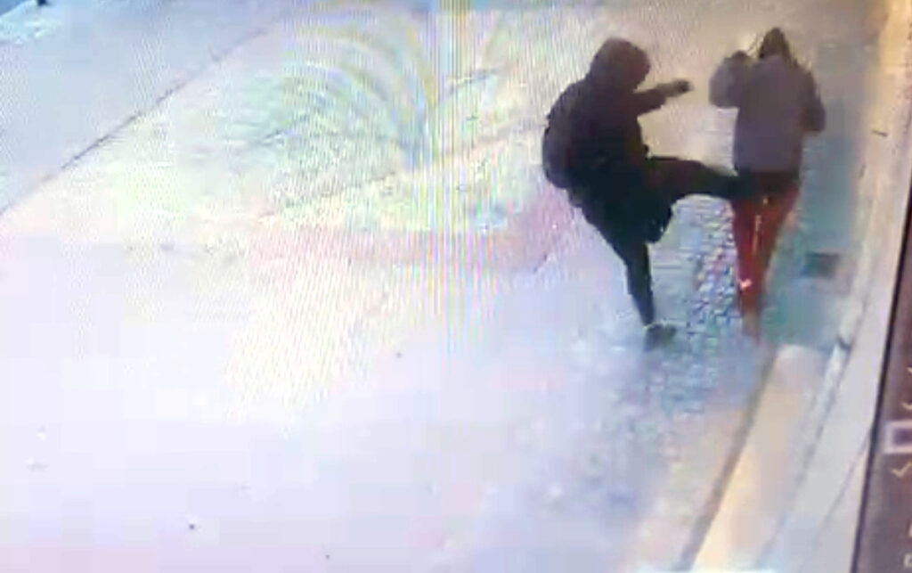 Mężczyzna w czarnej odzieży kopie inną osobę w jasnej kurtce na mokrej ulicy.