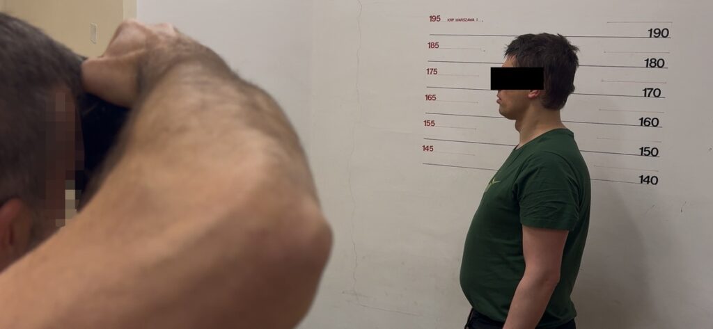 Mężczyzna w zielonej koszulce stoi na tle ściany z oznaczeniami wysokości, podczas gdy inna osoba go fotografuje.