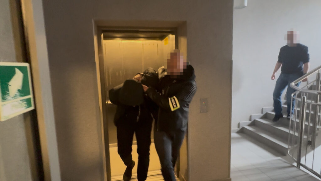 Mężczyzna w czarnej odzieży jest prowadzony przez drugiego mężczyznę w kurtce, w pobliżu windy w budynku.