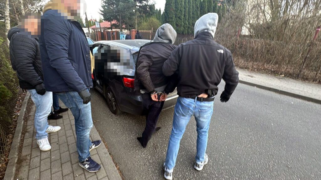 Grupa mężczyzn w odzieży roboczej zatrzymuje osobę z kajdankami przy czarnym samochodzie na ulicy.