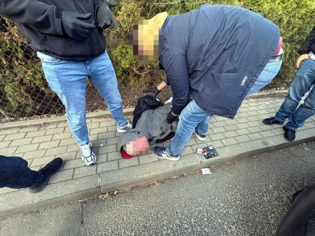 Grupa mężczyzn pomaga leżącemu na chodniku, jeden z nich przeszukuje jego plecak.